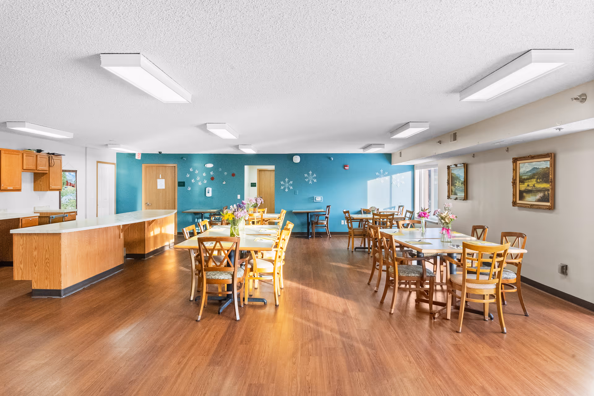 A bright and spacious dining room with wooden floors and several tables and chairs arranged neatly. The room has a teal accent wall with decorative snowflakes and handprints, wooden cabinets and a countertop on the left side, and framed landscape paintings on the right wall. Sunlight streams in through large windows, illuminating the space.