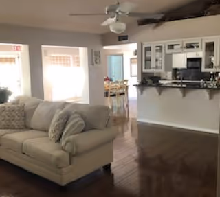 Open-plan interior showing a beige sofa on hardwood floors, ceiling fan, and a white kitchen with a breakfast bar in the background.