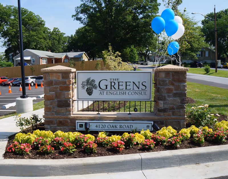 Stone sign at the entrance of The Greens at English Consul facility with decorative flowers planted in front and blue and white balloons tied to the right pillar. A parking lot and residential houses are visible in the background.