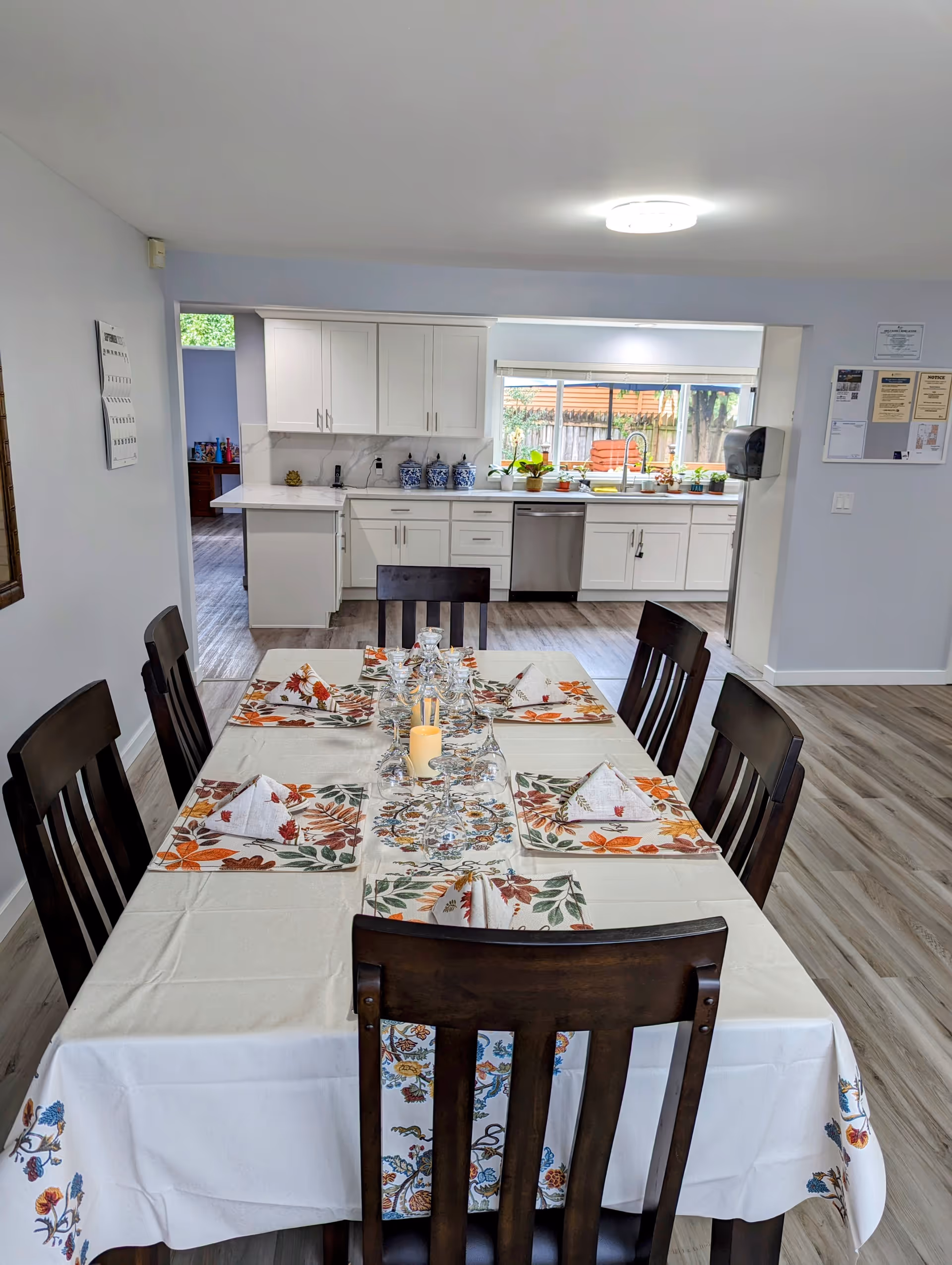 A dining area with a rectangular table covered with a white tablecloth decorated with floral patterns. The table is set with folded napkins, upside-down wine glasses, and a candle centerpiece. In the background, there is a kitchen with white cabinets, a dishwasher, a sink, and a window showing an outdoor view with plants.