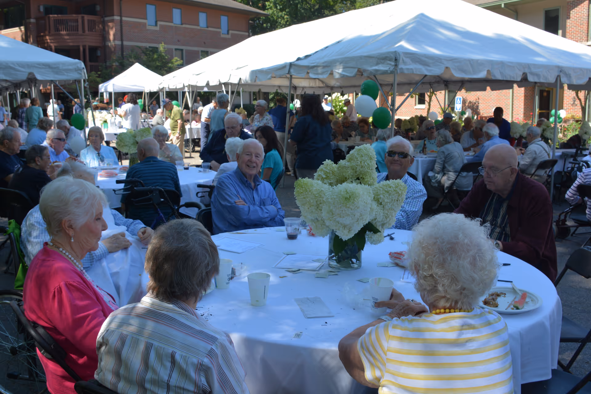 A group of elderly people sitting around a large round table covered with a white tablecloth, outdoors under a white tent. The table has a centerpiece of white flowers in a vase. Other tables with people are visible in the background, along with brick buildings and greenery.