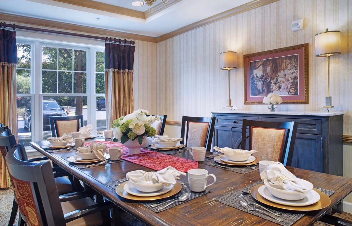 A dining room with a large wooden table set for six people, featuring white plates, bowls, cups, and napkins with gold napkin rings. A red table runner and a floral centerpiece decorate the table. The room has beige wallpaper, a large window with striped curtains, a dark wooden sideboard with two lamps, and a framed painting on the wall.