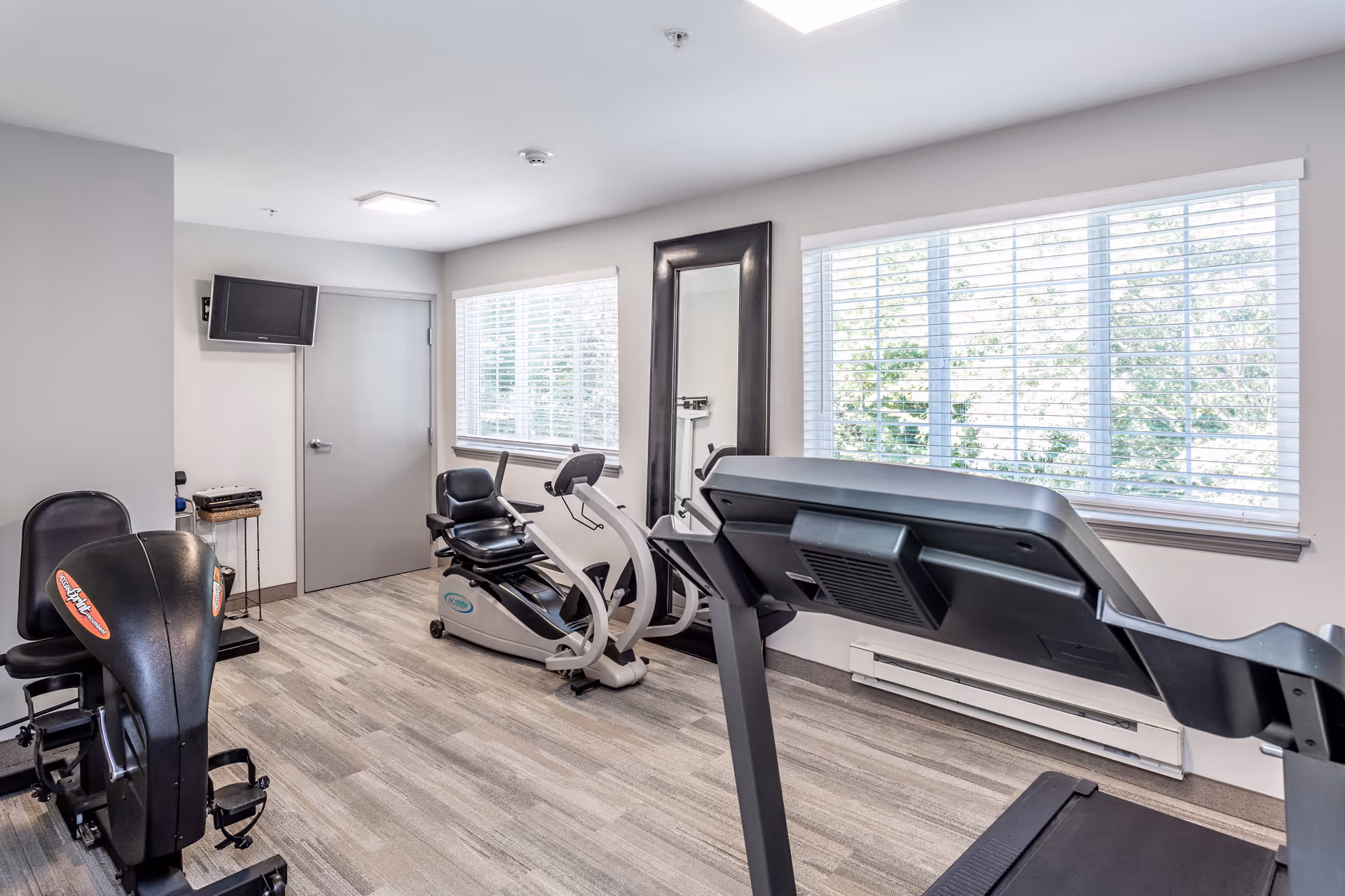 Small fitness room with a treadmill and recumbent bikes, large windows with blinds, and a wall mirror.