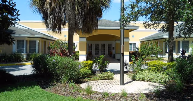 Exterior view of a single-story senior living facility building with a covered entrance, surrounded by landscaped greenery including bushes, trees, and a palm tree in the foreground.