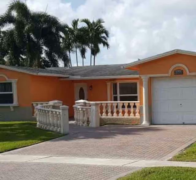 Front exterior view of a single-story orange house with a white garage door, a small porch with white balustrades, and palm trees in the background under a cloudy sky.