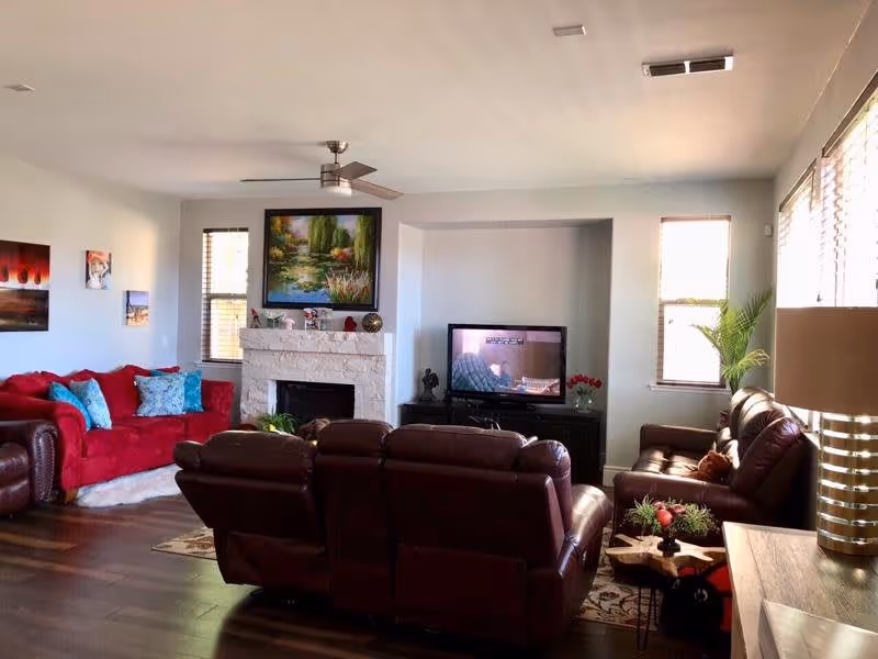A cozy living room with a red sofa adorned with blue and patterned pillows on the left, two brown leather recliners in the center facing a TV, and a brown leather sofa on the right. The room features a white stone fireplace with decorative items and a large painting above it. There are windows with blinds on either side of the fireplace and a ceiling fan above. A lamp and a small table with plants are visible on the right side.