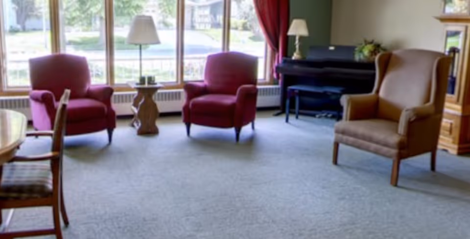 Sunlit common living area with armchairs, side tables, a piano, and large windows overlooking greenery.