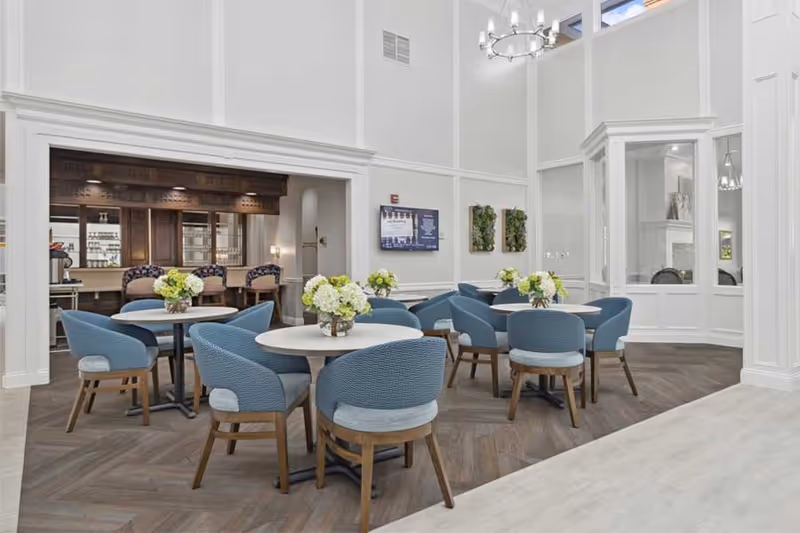 A bright and elegant dining area with round tables and blue upholstered chairs. Each table has a floral centerpiece. The room features high ceilings, white walls with decorative molding, a chandelier, and a fireplace visible through glass panels. In the background, there is a bar area with wooden cabinetry and bar stools.