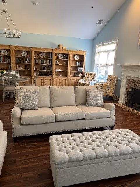 A cozy living room area with a beige three-seat sofa adorned with two patterned cushions, a matching tufted ottoman in front, wooden bookshelves filled with books and decorative plates along the back wall, two floral armchairs near a large window letting in natural light, and a fireplace on the right side.