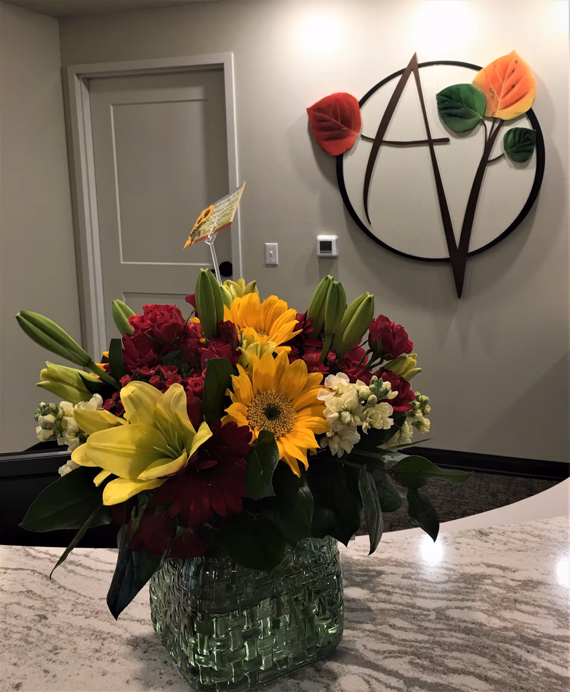 A colorful bouquet of sunflowers and lilies in a green glass vase sits on a marble countertop with a decorative wall logo and door in the reception area behind it.