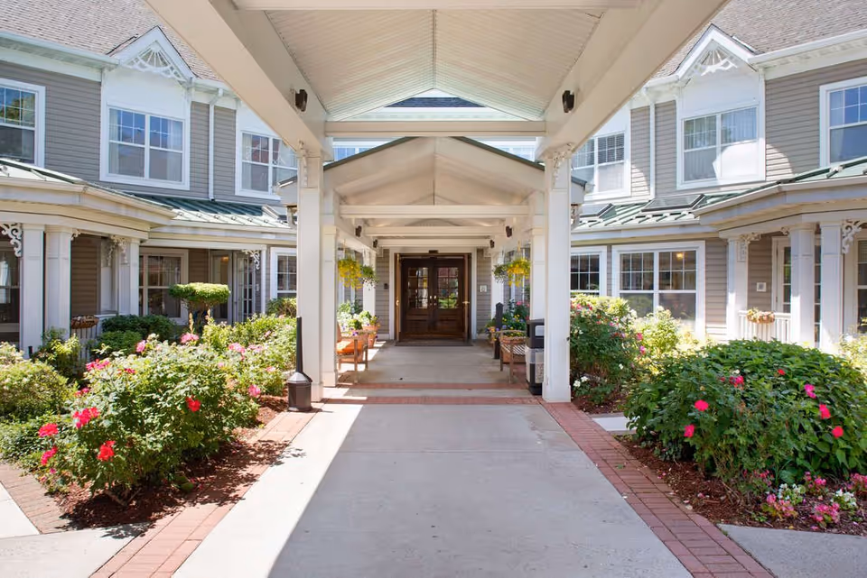 Covered entrance walkway leading to the main doors of a two-story senior living building flanked by flowerbeds and benches.