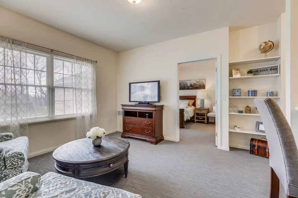 A bright living room with a large window covered by sheer curtains, two patterned armchairs, and a round wooden coffee table with a vase of white flowers. A wooden TV stand with a flat-screen TV is against the wall. An open doorway leads to a bedroom with a bed, nightstand, and lamp. To the right, built-in shelves hold decorative items and a gray upholstered chair is partially visible.