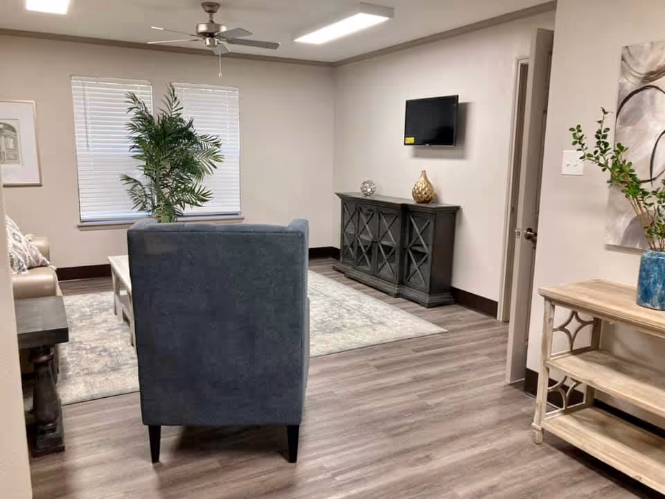 A cozy living room with a gray armchair facing a wall-mounted TV above a dark wooden cabinet. There is a large window with blinds, a ceiling fan, a light-colored rug, a beige sofa, a coffee table, and decorative plants and artwork on the walls.
