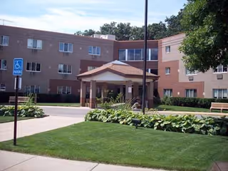 Exterior view of a multi-story brick nursing center building with a covered entrance, surrounded by green lawns, plants, and a tree. A handicapped parking sign is visible near the driveway.