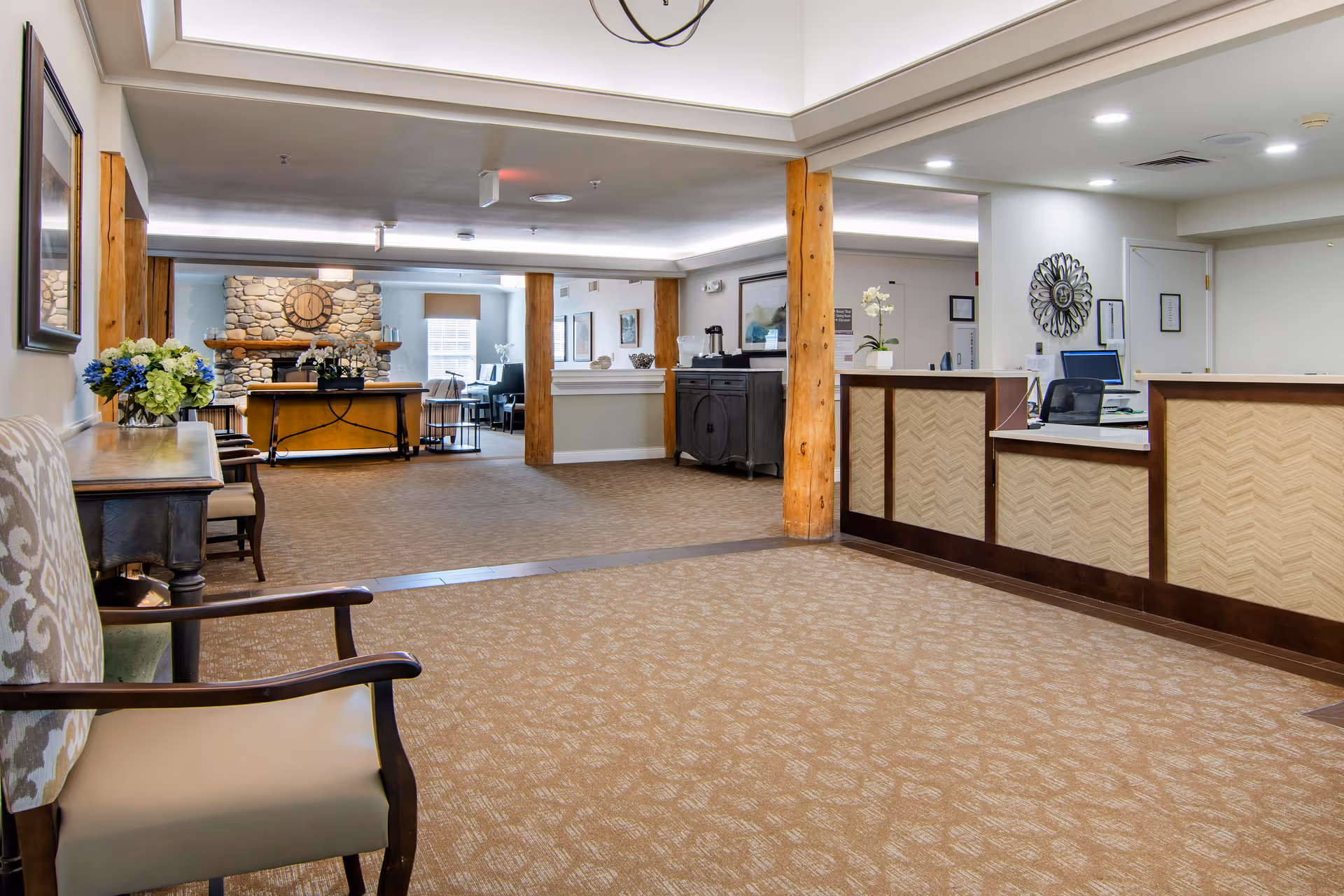 A spacious senior living facility lobby with a reception desk on the right, wooden support columns, a seating area with chairs and a table with flowers on the left, and a stone fireplace with a large clock in the background.