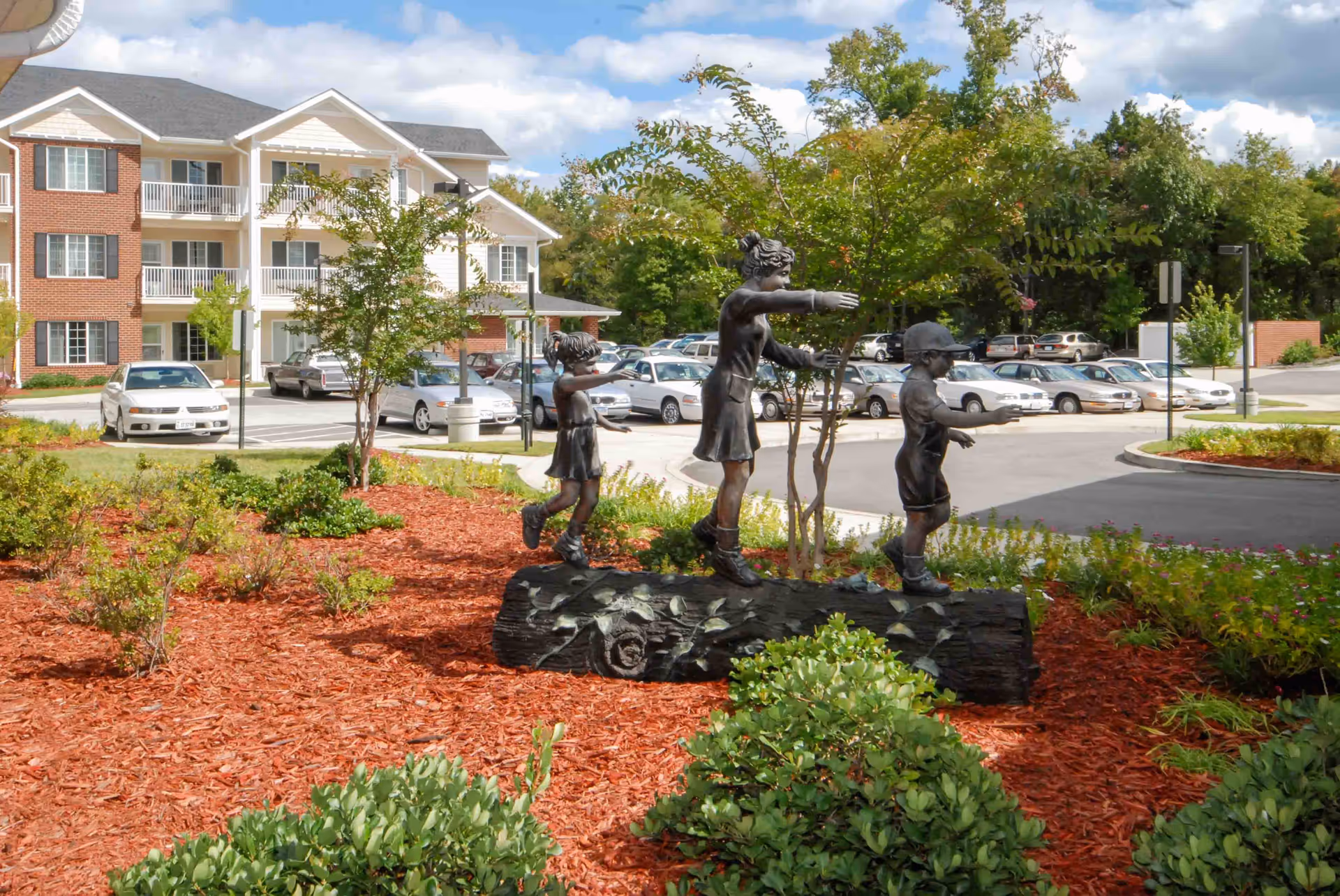 Outdoor garden area with a bronze statue of three children playing on a log surrounded by mulch and greenery, with a parking lot and a multi-story residential building in the background under a partly cloudy sky.