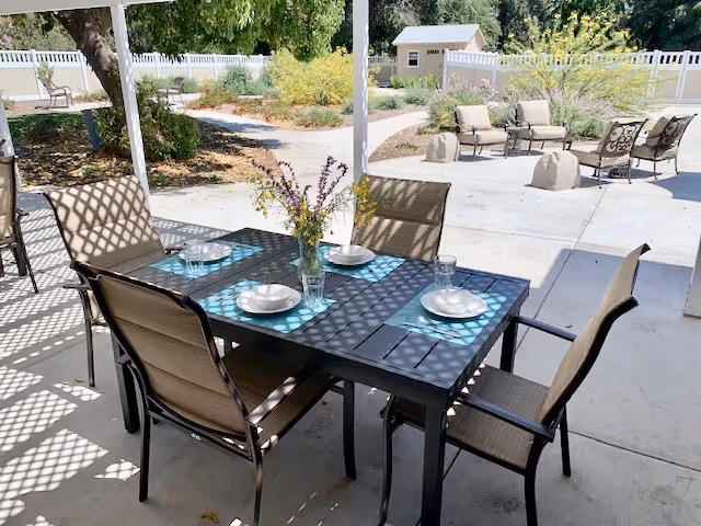 Outdoor patio area with a black metal table set for four with plates, glasses, and a vase of flowers. Surrounding the patio are chairs, a white fence, trees, and garden landscaping with a small shed in the background.
