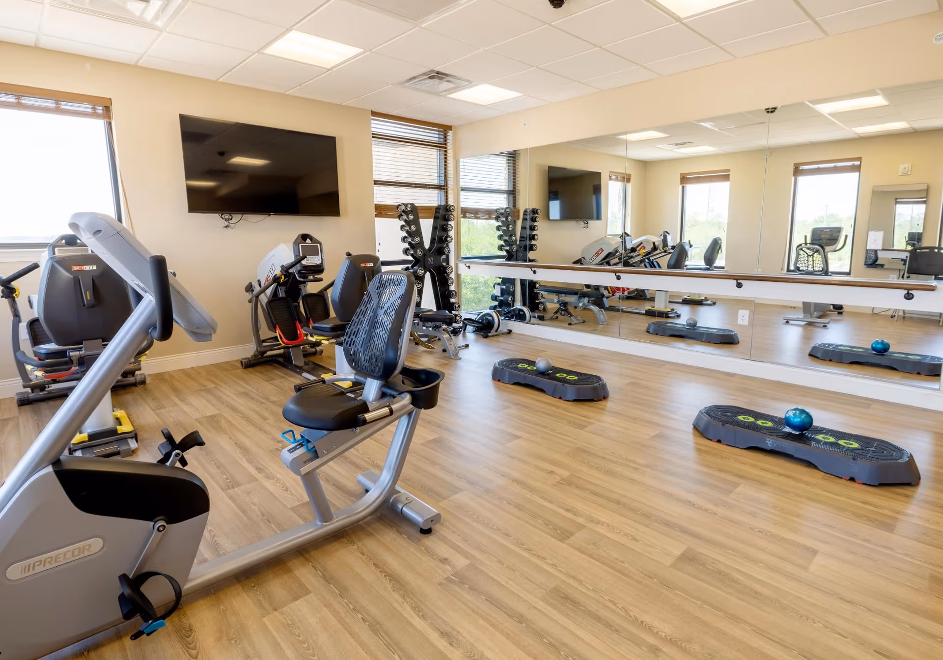 Bright fitness room with recumbent exercise bikes, step platforms, free weights, a wall mirror and a mounted TV.