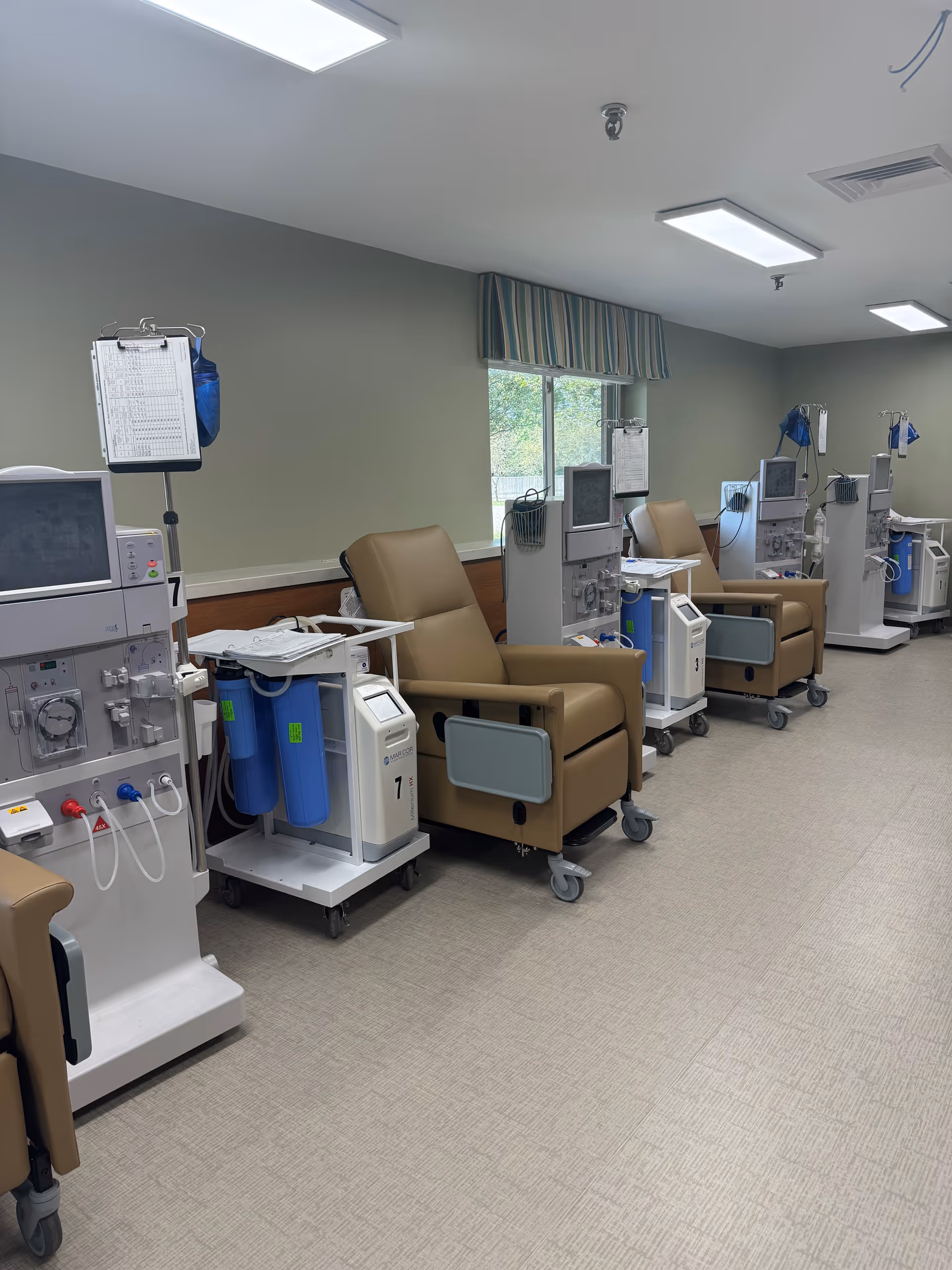 A medical treatment room with several beige recliner chairs lined up against a wall, each paired with dialysis machines and IV stands. The room has a window with a striped valance, overhead fluorescent lighting, and a light-colored floor.