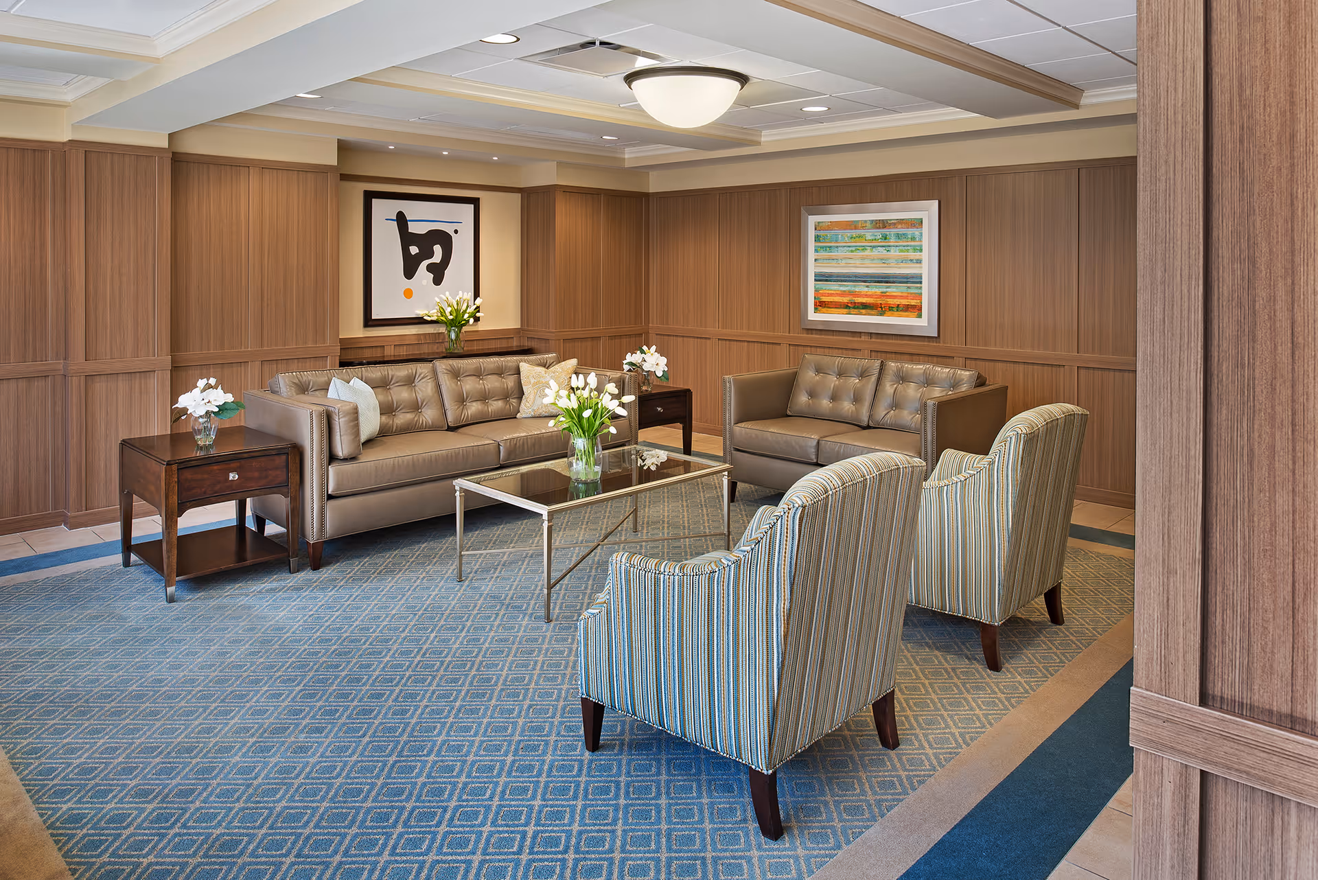 A cozy living room area with two brown leather sofas, two striped armchairs, a glass coffee table, and two wooden side tables with flower vases. The walls are paneled with wood, and there are two framed artworks hanging on the walls. The floor is covered with a blue patterned carpet.