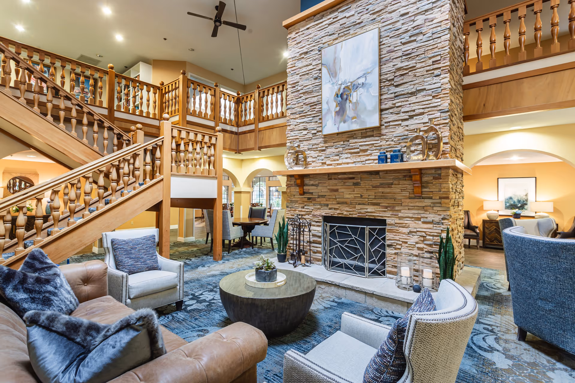 A spacious living room area in a senior living facility featuring a large stone fireplace with modern artwork above it. The room has comfortable seating including a leather sofa, armchairs with cushions, and a round coffee table. There is a wooden staircase with decorative railings leading to an upper level balcony. The space is warmly lit with a mix of natural and artificial light, and the decor includes plants and candles.