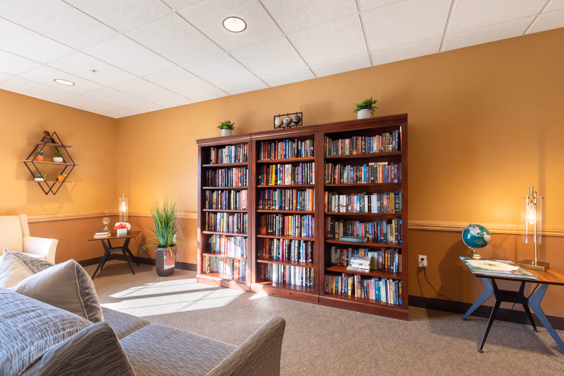 A cozy reading room with a large wooden bookshelf filled with books against a warm beige wall. The room features a comfortable sofa with pillows, a small side table with a lamp and a globe, a potted plant, and decorative wall shelves with small plants and ornaments. Sunlight streams in, creating a welcoming atmosphere.