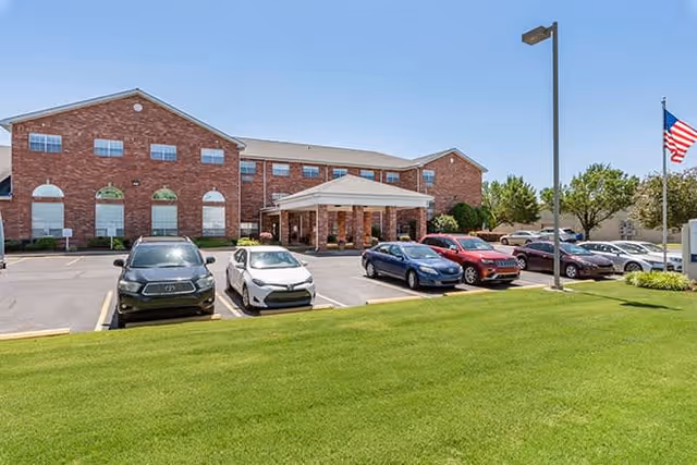 Front exterior view of a two-story brick building with a covered entrance, several parked cars in the parking lot, a green lawn in the foreground, a street lamp, and an American flag on a flagpole to the right.