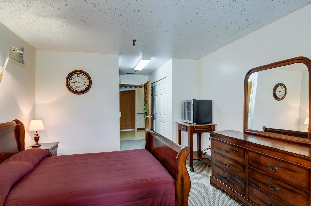 Furnished bedroom with a wooden sleigh bed, matching dresser and mirror, wall clock, small TV on a stand, and an open doorway.