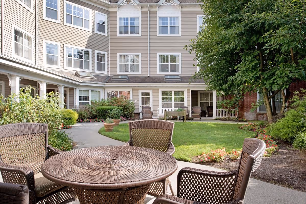 Outdoor courtyard area at Sunrise at Fleetwood featuring a round wicker table with four matching chairs in the foreground, surrounded by a well-maintained lawn, flower beds, shrubs, and trees. The multi-story building with beige siding and multiple windows encloses the courtyard.