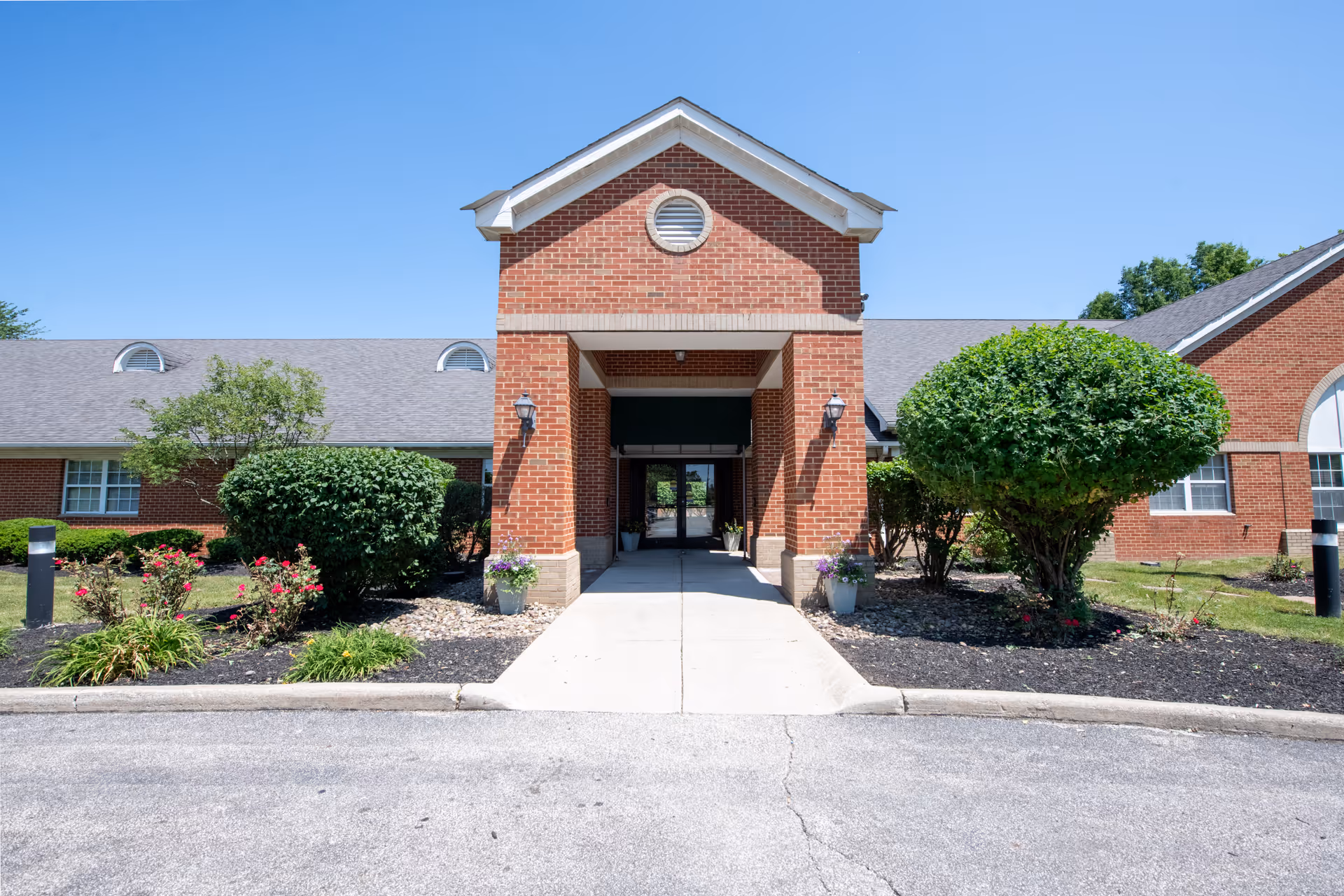 Front exterior view of Hopkins Rehabilitation and Care Center showing a brick building with a covered entrance, well-maintained landscaping including bushes and flowers, and a clear blue sky.