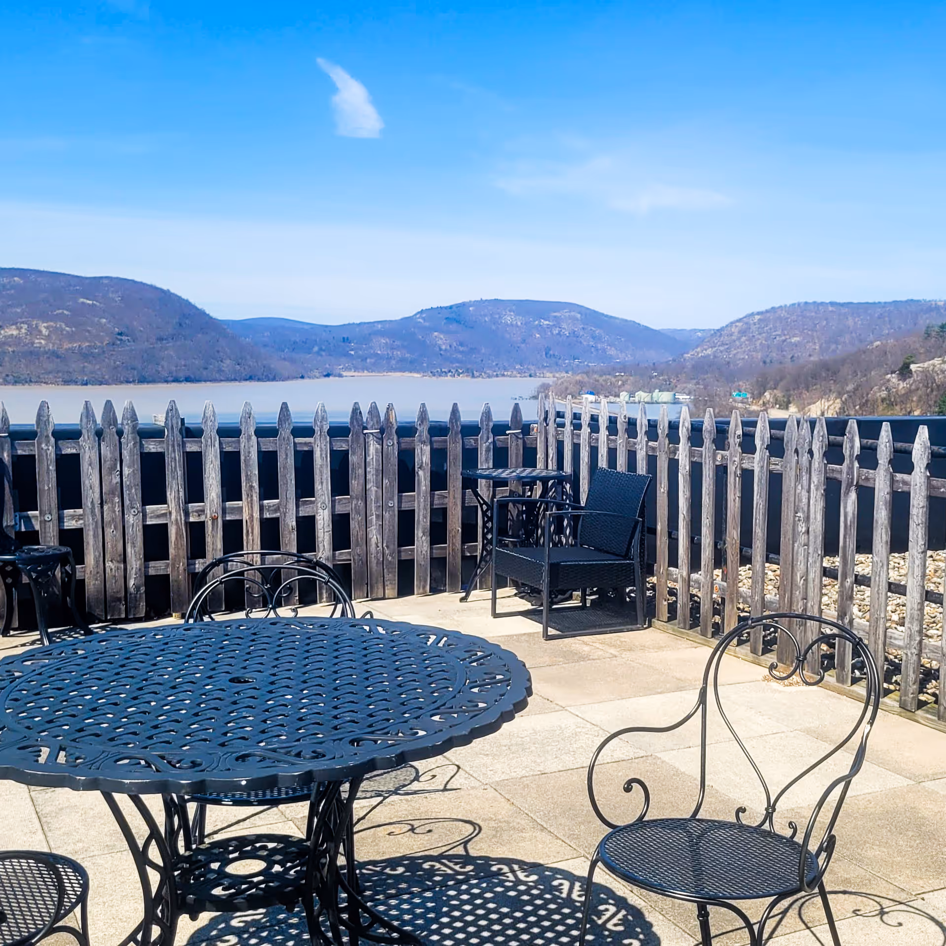 Outdoor patio area with black wrought iron tables and chairs, surrounded by a wooden picket fence. In the background, there is a scenic view of a river and forested hills under a clear blue sky.