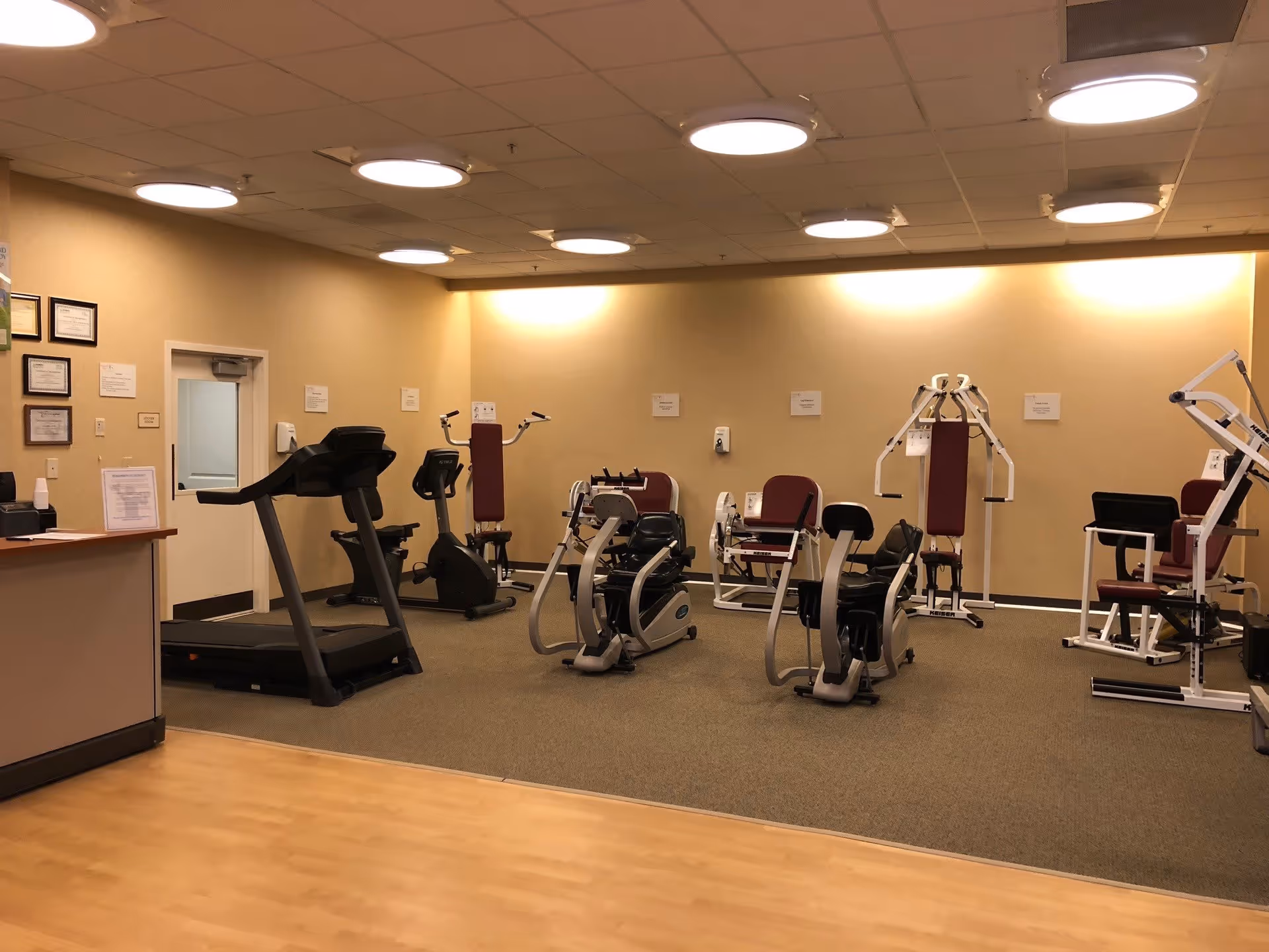 Interior view of a fitness room in a senior living facility with various exercise machines including treadmills, stationary bikes, and weight machines. The room has beige walls, a carpeted exercise area, and a wooden floor section near the entrance. Several certificates and notices are displayed on the wall near a door.