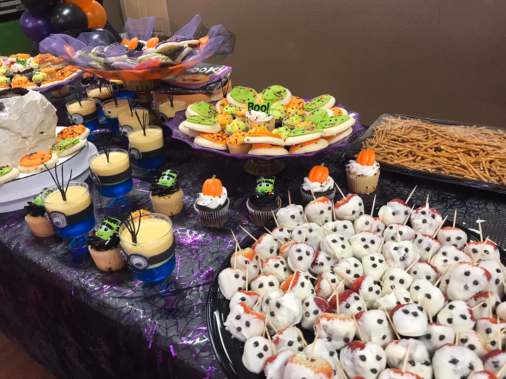 A Halloween-themed dessert table with cupcakes, decorated cookies, dipped strawberries, pretzels, and layered pudding cups.