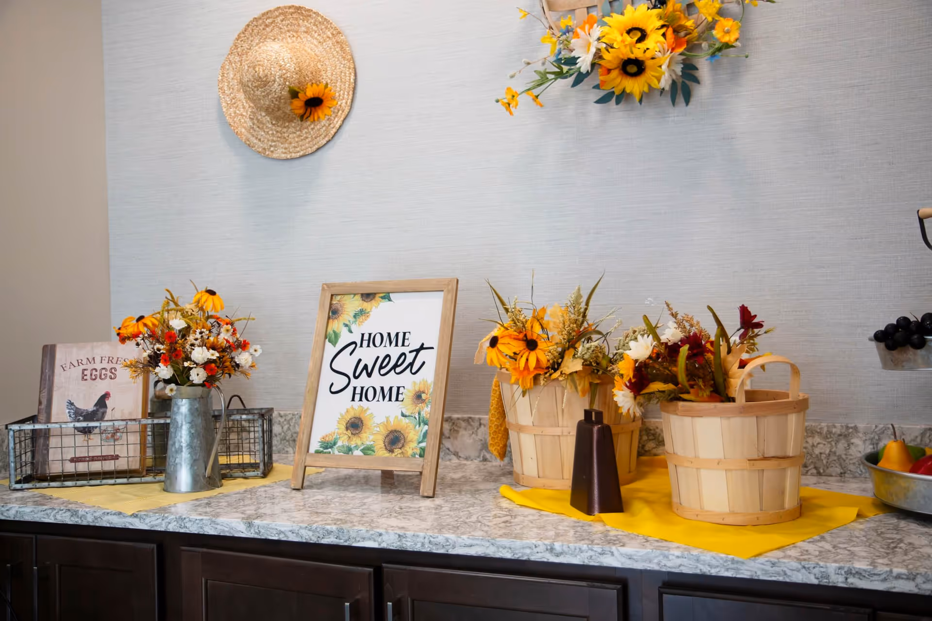A countertop decorated with a framed sign that says 'Home Sweet Home' surrounded by sunflower-themed floral arrangements in metal and wooden containers. A straw hat with a sunflower is hanging on the wall above, and a small sign reading 'Farm Fresh Eggs' with a chicken illustration is also on the counter.