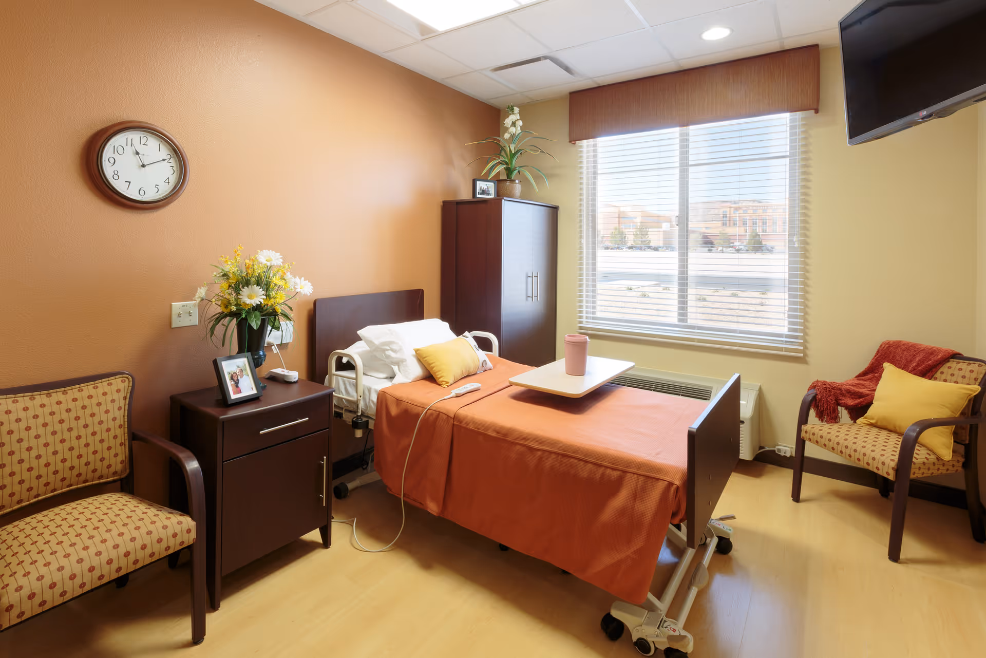 A well-lit patient room with an adjustable hospital bed covered in an orange blanket, a small overbed table with a pink cup, two patterned armchairs, a wooden nightstand with a vase of flowers and a framed photo, a wall clock, a large window with blinds, and a wall-mounted TV.