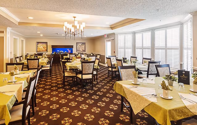 Dining room with multiple tables set with yellow tablecloths and chairs, large shuttered windows, a chandelier, and an aquarium at the far wall.