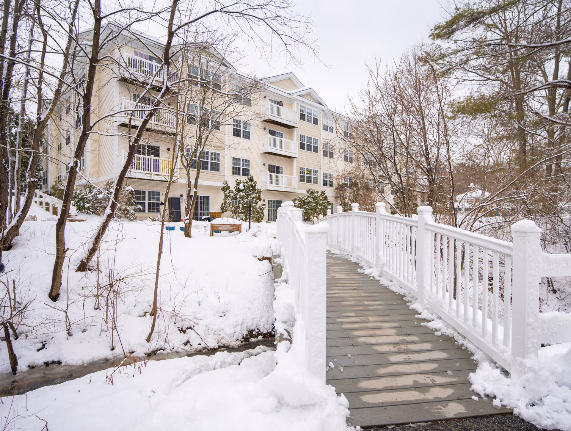Snow-covered outdoor scene featuring a wooden footbridge with white railings leading towards a multi-story residential building with balconies. Bare trees and snow-covered ground surround the area.