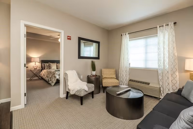 A cozy living room with a dark gray sofa, a round black coffee table with a remote control on it, a patterned armchair, and a small side table with a plant. A window with white curtains lets in natural light. Through an open doorway, a bedroom with a bed, pillows, and a bedside lamp is visible.