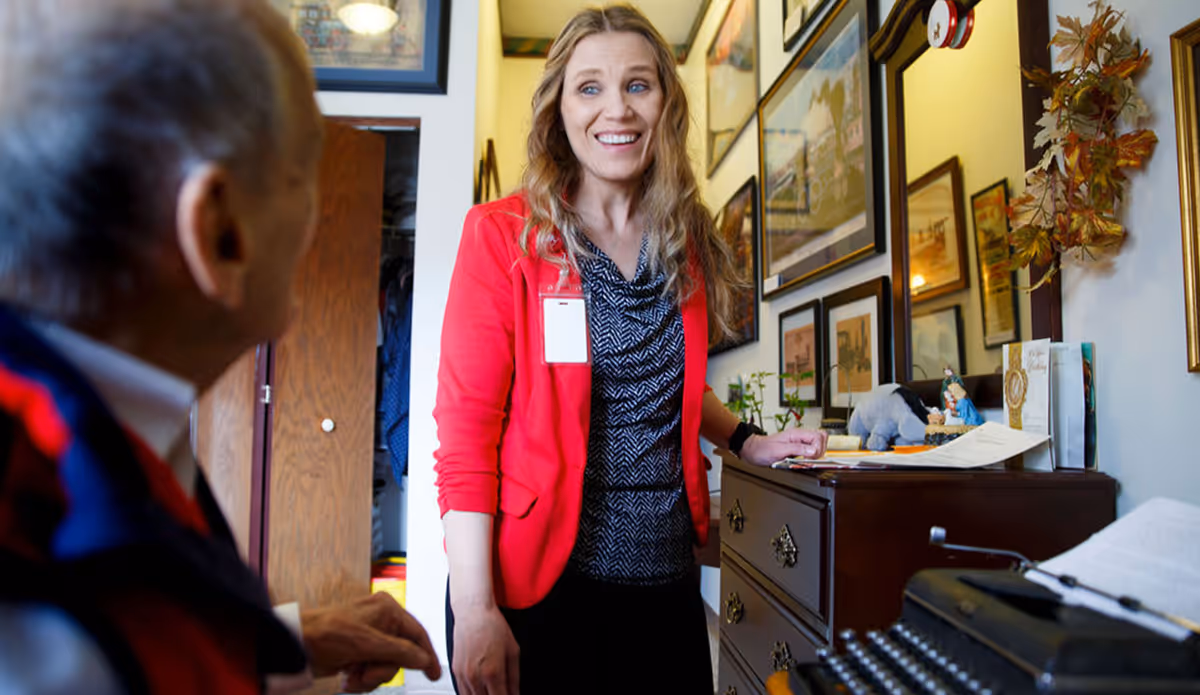 A woman in a red blazer with a name badge is smiling and standing next to a dresser in a room decorated with framed pictures on the wall. An elderly person is partially visible in the foreground, facing the woman.