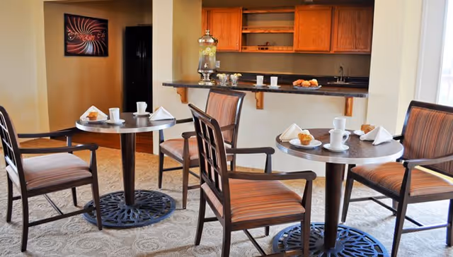 A cozy dining area with two round tables, each set with white cups, plates, napkins, and muffins. Four wooden chairs with striped cushions surround the tables. In the background, there is a kitchen counter with wooden cabinets, a beverage dispenser, and some pastries. The room has warm lighting and a carpeted floor.