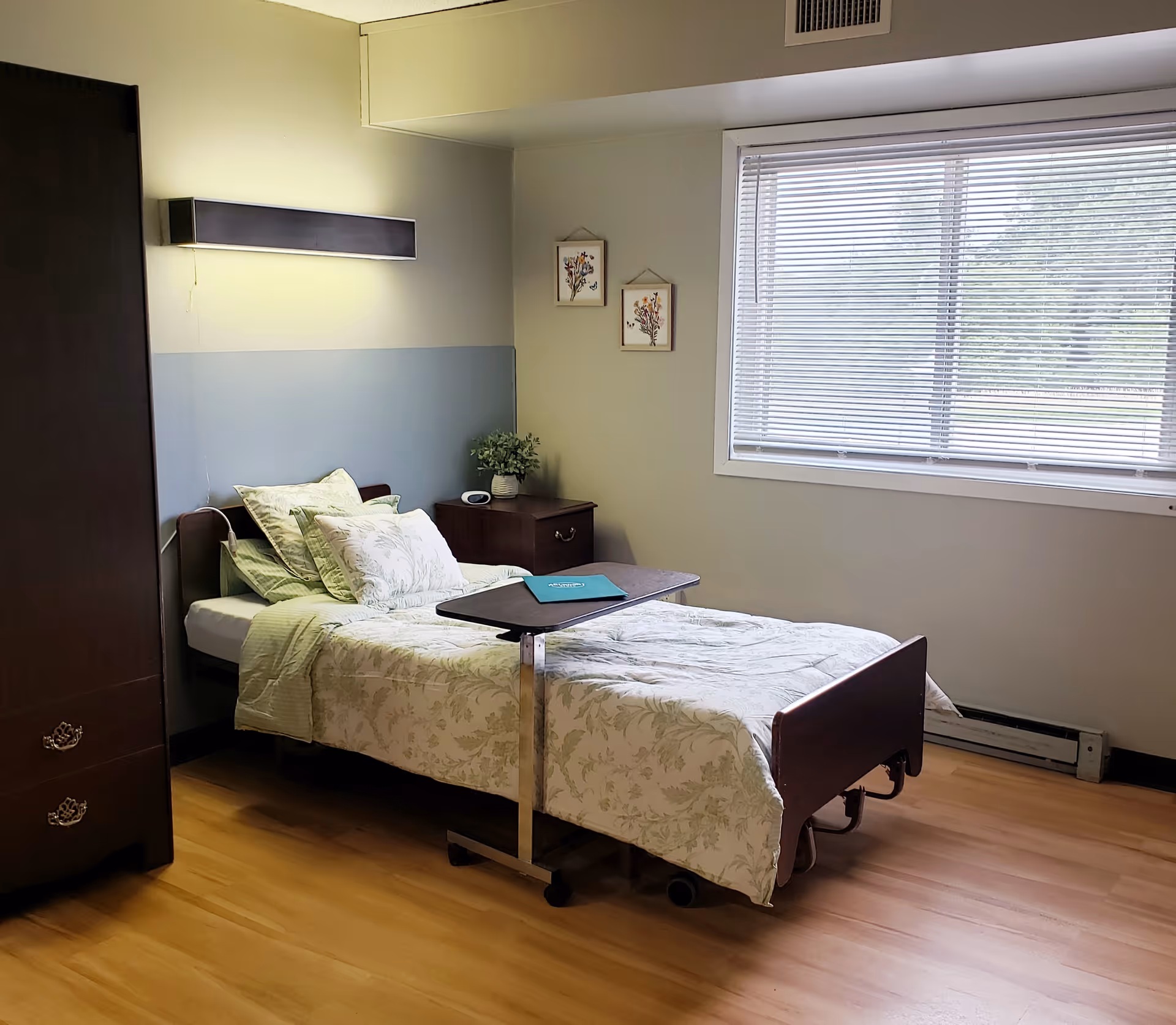 A single bed with floral bedding and multiple pillows in a well-lit room. The bed has an adjustable overbed table with a folder on it. Next to the bed is a wooden nightstand with a small plant and a clock. There is a large window with blinds letting in natural light, and two small framed floral pictures hang on the wall above the nightstand. A dark wooden wardrobe is partially visible on the left side of the image. The floor is wooden, and the walls are painted in light colors.