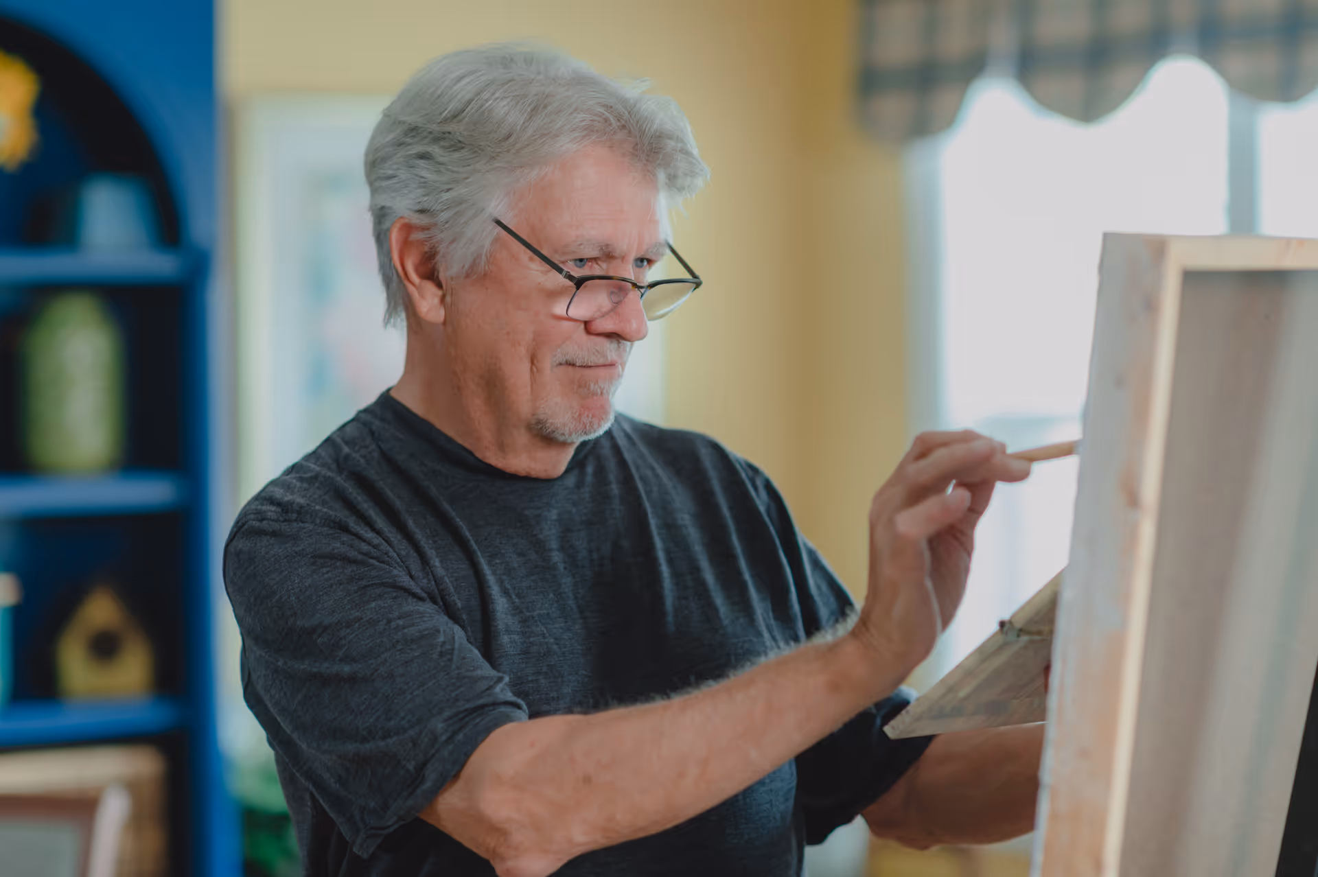 An elderly man with gray hair and glasses is painting on a canvas in a well-lit room with a blue shelf and framed artwork in the background.
