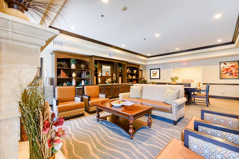 Bright furnished common lounge with sofas, armchairs, a wooden coffee table, built-in shelving and a dining area.