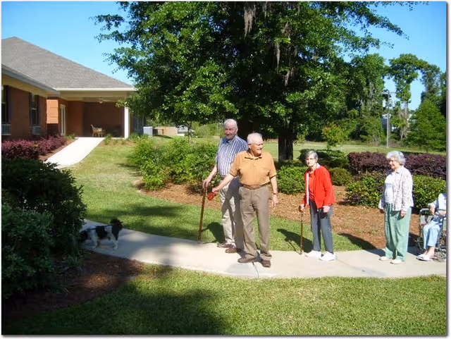 Four elderly people standing on a curved sidewalk in a garden area outside a building. Two men and two women are visible, with one man and one woman using canes. A small black and white dog is also on the sidewalk near some bushes. The scene is sunny with green grass, trees, and shrubs surrounding the area.
