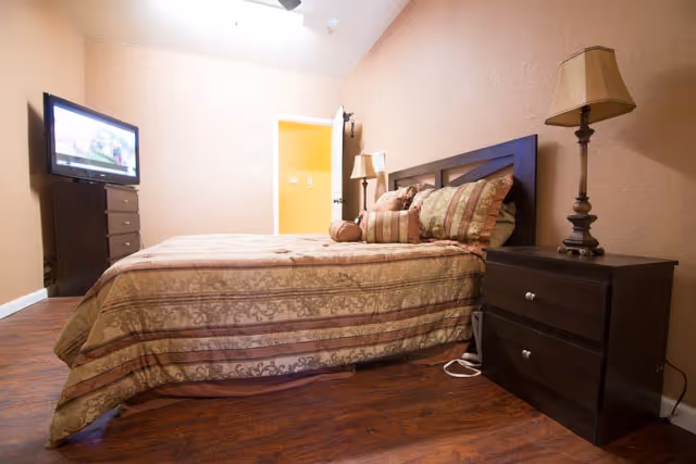 A bedroom with a double bed covered in a patterned beige and brown bedspread, two matching pillows, and a dark wooden headboard. There is a dark wooden nightstand with a lamp on the right side of the bed. A flat-screen TV is mounted on a dark wooden dresser against the left wall. The room has wooden flooring and peach-colored walls, with an open door leading to another room.