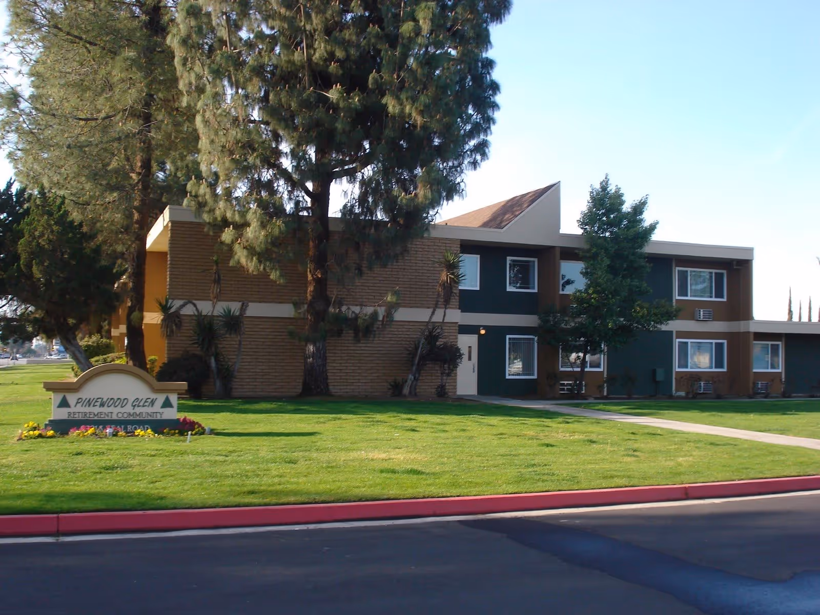 Exterior view of a two-story retirement community building with green and brown walls, surrounded by trees and a well-maintained lawn. A sign in front reads 'Pinewood Glen Retirement Community'.