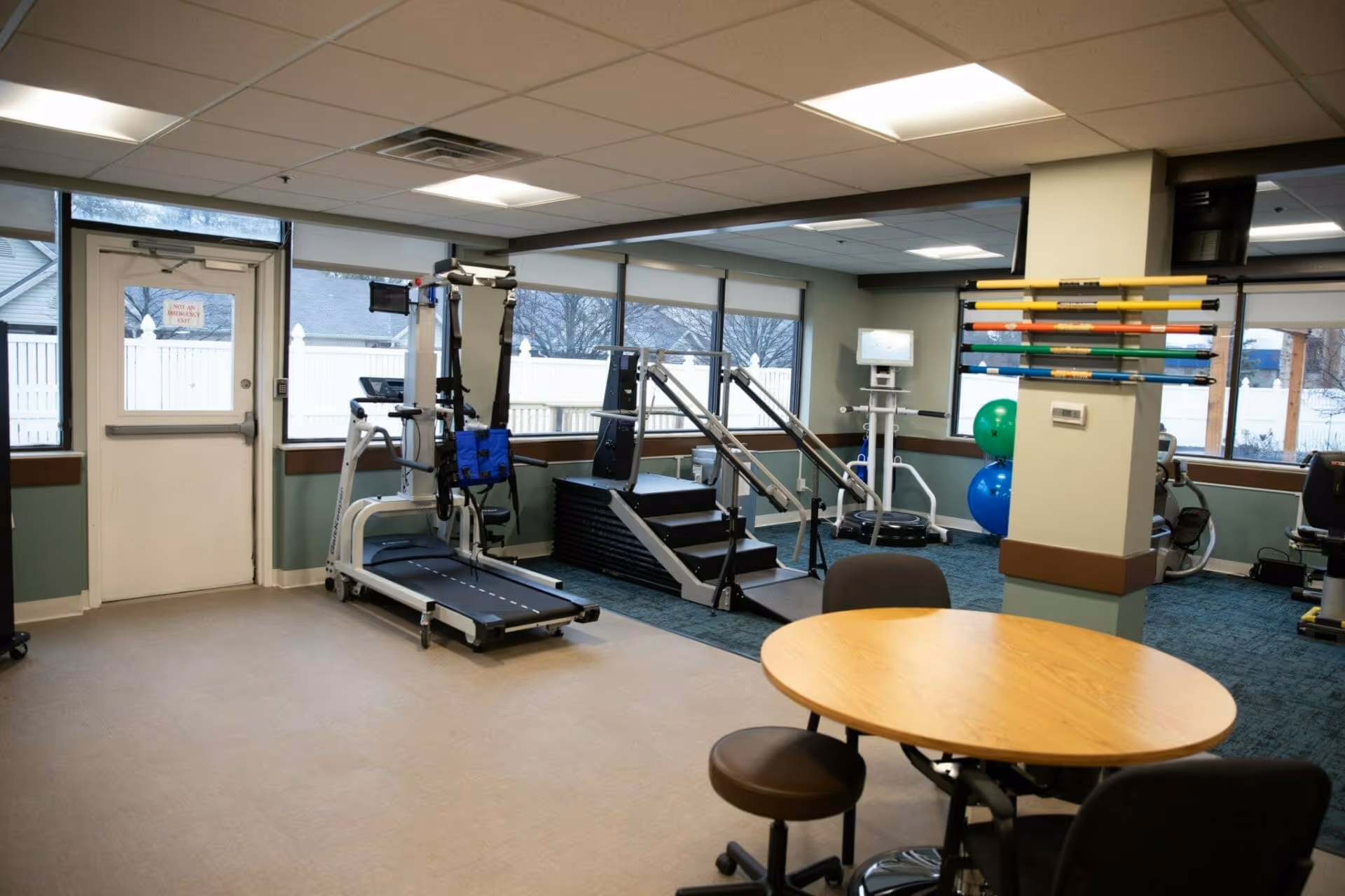 Interior view of a rehabilitation or physical therapy room with exercise equipment including a treadmill, stairs with handrails, therapy balls, and a rack of colorful exercise bars. There is a round wooden table with chairs in the foreground and large windows letting in natural light.