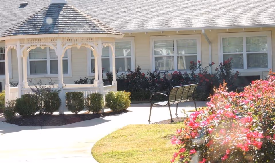 Outdoor garden area at Iris Memory Care of Nichols Hills featuring a white gazebo, a black metal bench, green bushes, and blooming pink flowers in front of a beige building with multiple windows.