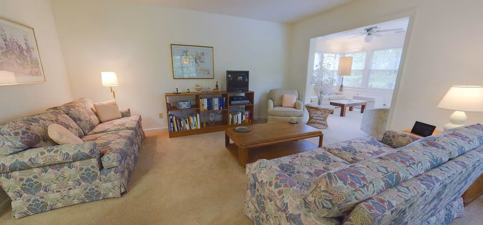 A living room with two floral patterned sofas facing a wooden coffee table. There is a wooden bookshelf against the wall with books, a small TV, and decorative items. A beige armchair with a pink pillow is next to the bookshelf. The room opens into a sunroom with large windows, a ceiling fan, a floor lamp, and additional seating including a wicker table and chairs.