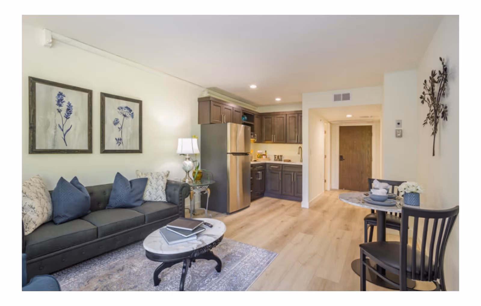 A cozy living area with a dark gray sofa adorned with blue and patterned pillows, a marble coffee table with books on it, and two framed botanical prints on the wall. To the right, there is a small round dining table set with two black chairs and tableware. In the background, a kitchen area with dark wood cabinets, a stainless steel refrigerator, and a countertop with a decorative 'EAT' sign is visible. The floor is light wood, and the walls are painted white.